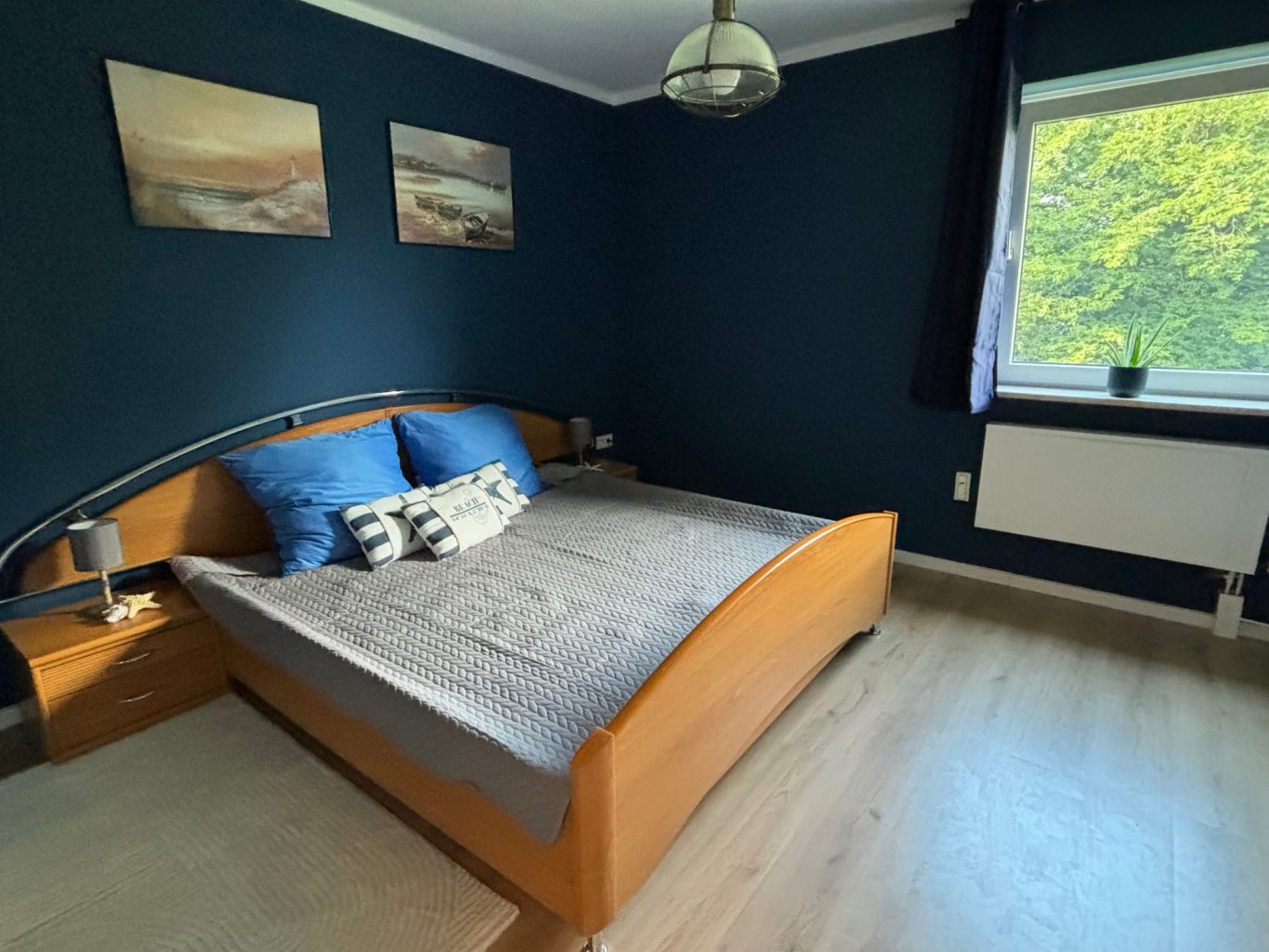 Schlafzimmer mit blauer Wandfarbe, Bett, zwei Kissen und Fenster mit Pflanzen.