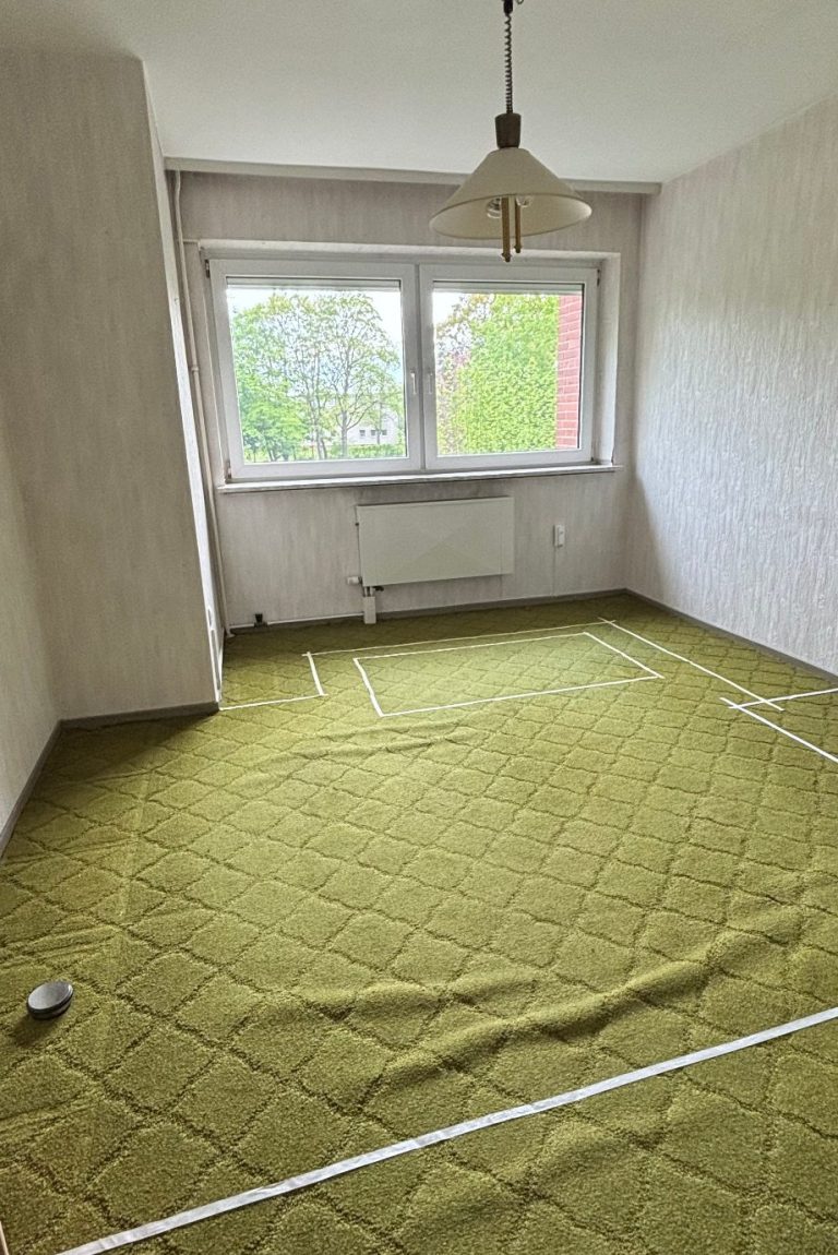 Leerer Raum mit grünem Teppich, Fenster und einer alten Lampe an der Decke.